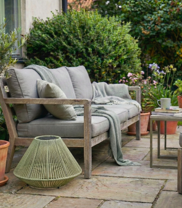 Un confortable banc en bois, agrémenté d’un coussin et d’une couverture, prend place sur une terrasse de jardin entourée de plantes et accompagné d’une petite table d’appoint.