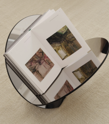Table d’appoint ronde en verre foncé avec un livre d’art ouvert, sur un tapis beige.