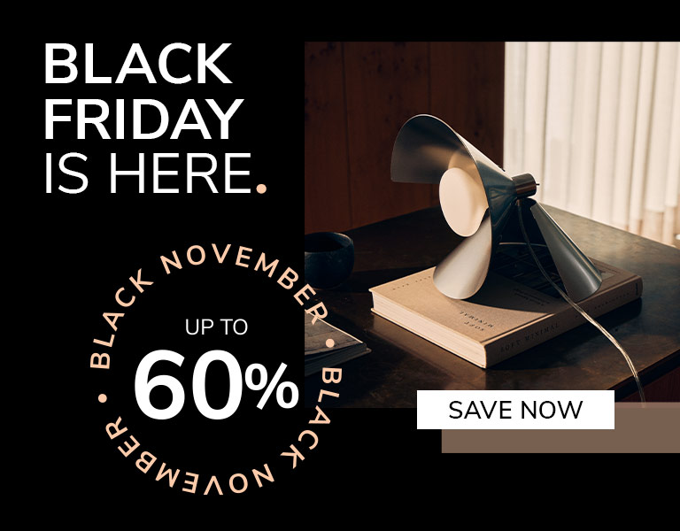 A modern table lamp with angled metal blades sits on top of a book on a dark wooden desk in soft daylight. Text reads: ‘Black Friday is here. Up to 60%. Save Now.
